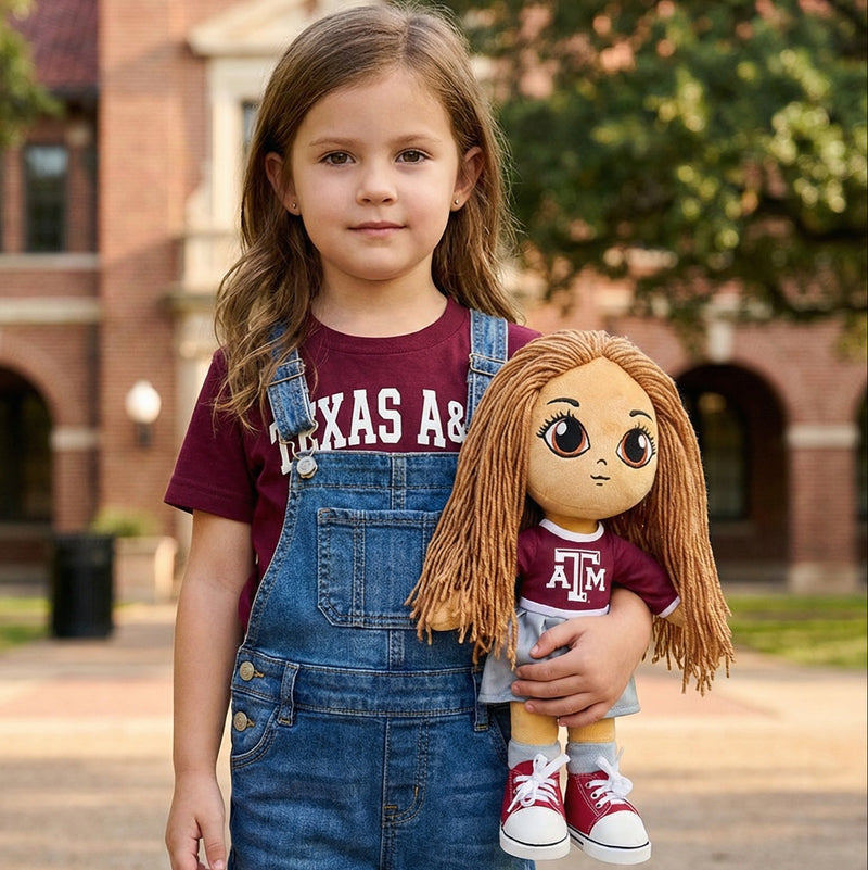 Young girl holding a doll with Texas A&M University attire in front of a brick building.